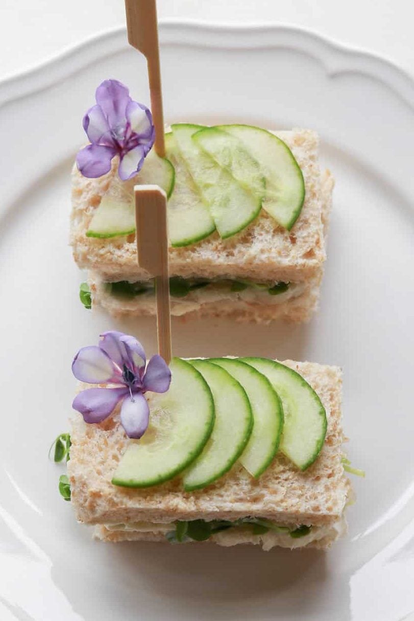 Two neatly stacked cucumber tea sandwiches on a white plate, each secured with a wooden stick. The sandwiches are garnished with thin slices of cucumber on top and small purple flowers for decoration.
