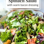 A bowl of spinach salad topped with bacon pieces, shaved cheese, and a flavorful warm bacon dressing, with salad tongs beside the bowl.