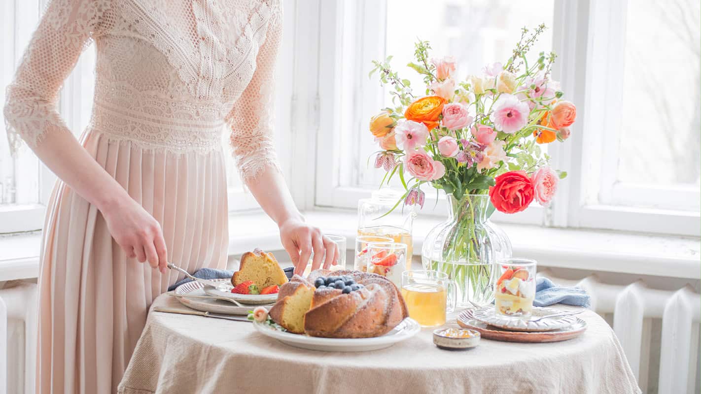 A person in a light pink lace dress sets a table for afternoon tea with cake, fruit, drinks, and a vase of colorful flowers near a bright window, showing how to serve full-size desserts with elegance.
