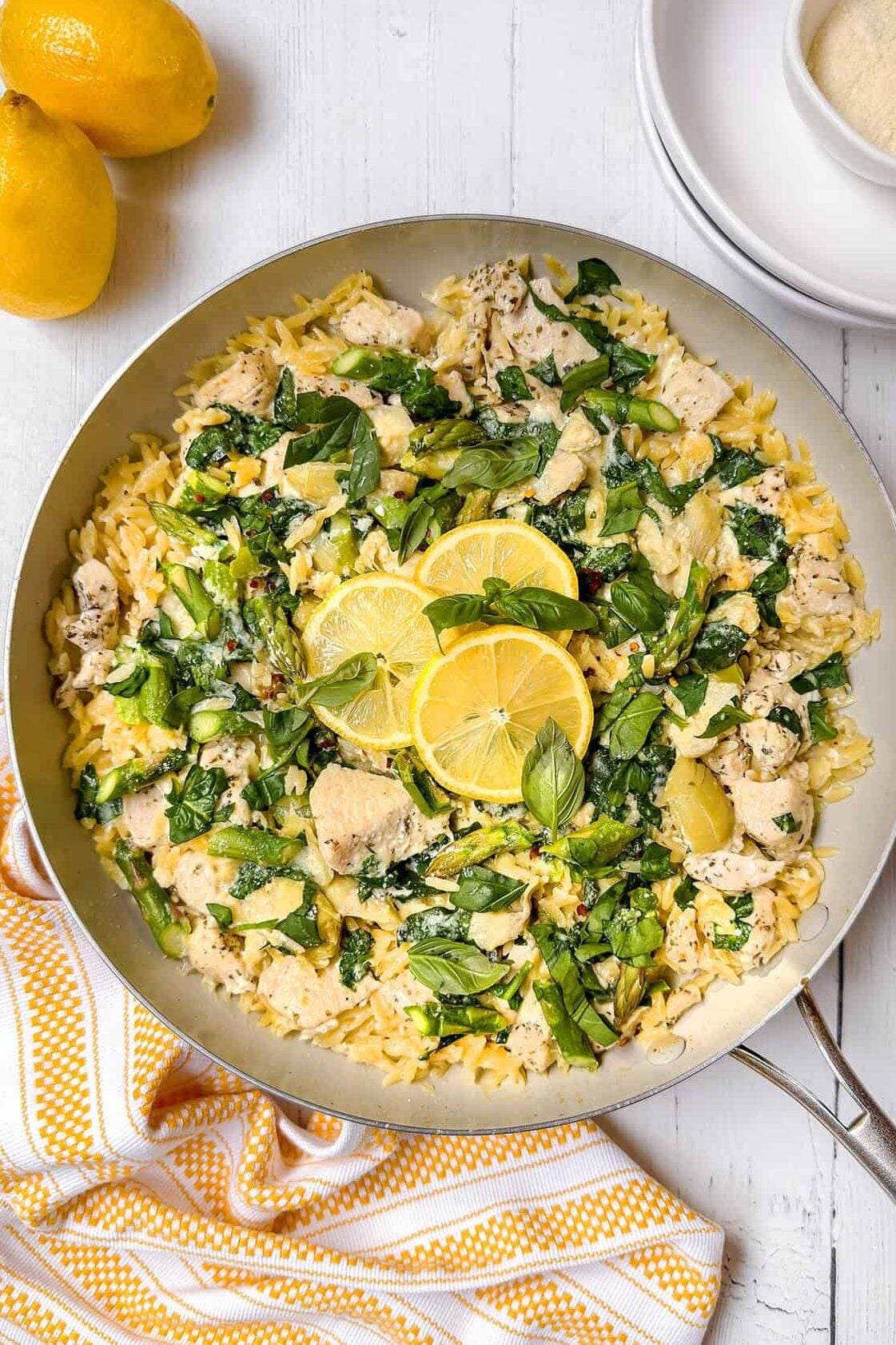 A skillet filled with lemon chicken orzo, spinach, and topped with lemon slices, served on a white wooden surface with fresh lemons, Parmesan cheese, and a yellow-striped towel nearby.