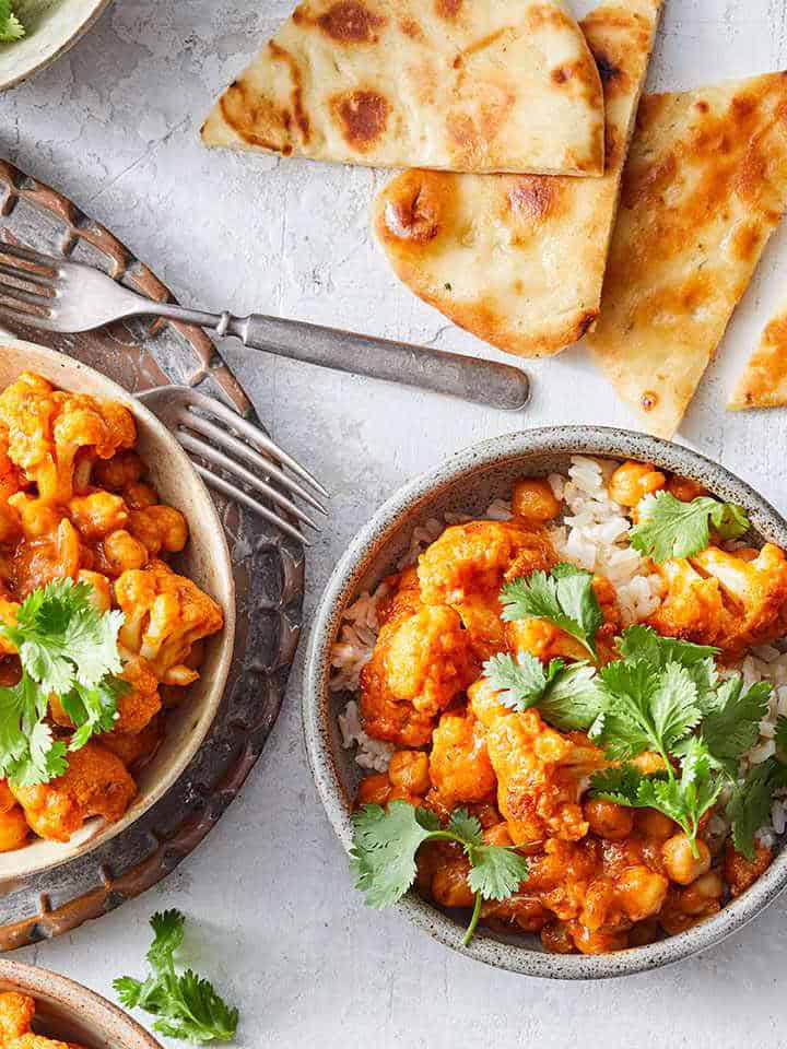 Two bowls of rice topped with a chickpea and cauliflower curry, garnished with cilantro, are served alongside naan bread-a flavorful option from the April 30 Minute Weeknight Dinners Meal Plan on a light-colored surface.