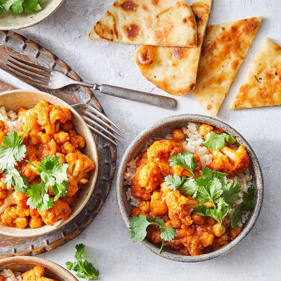Two bowls of rice topped with a chickpea and cauliflower curry, garnished with cilantro, are served alongside naan bread-a flavorful option from the April 30 Minute Weeknight Dinners Meal Plan on a light-colored surface.