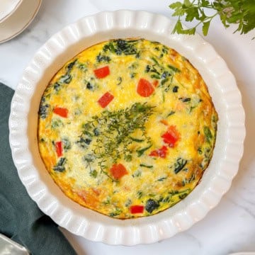 A baked vegetable frittata, perfect as a crustless quiche recipe, features red bell pepper pieces and leafy greens, garnished with fresh herbs, and sits in a white fluted dish on a marble surface.