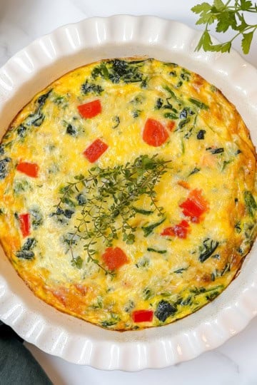 A baked vegetable frittata, perfect as a crustless quiche recipe, features red bell pepper pieces and leafy greens, garnished with fresh herbs, and sits in a white fluted dish on a marble surface.