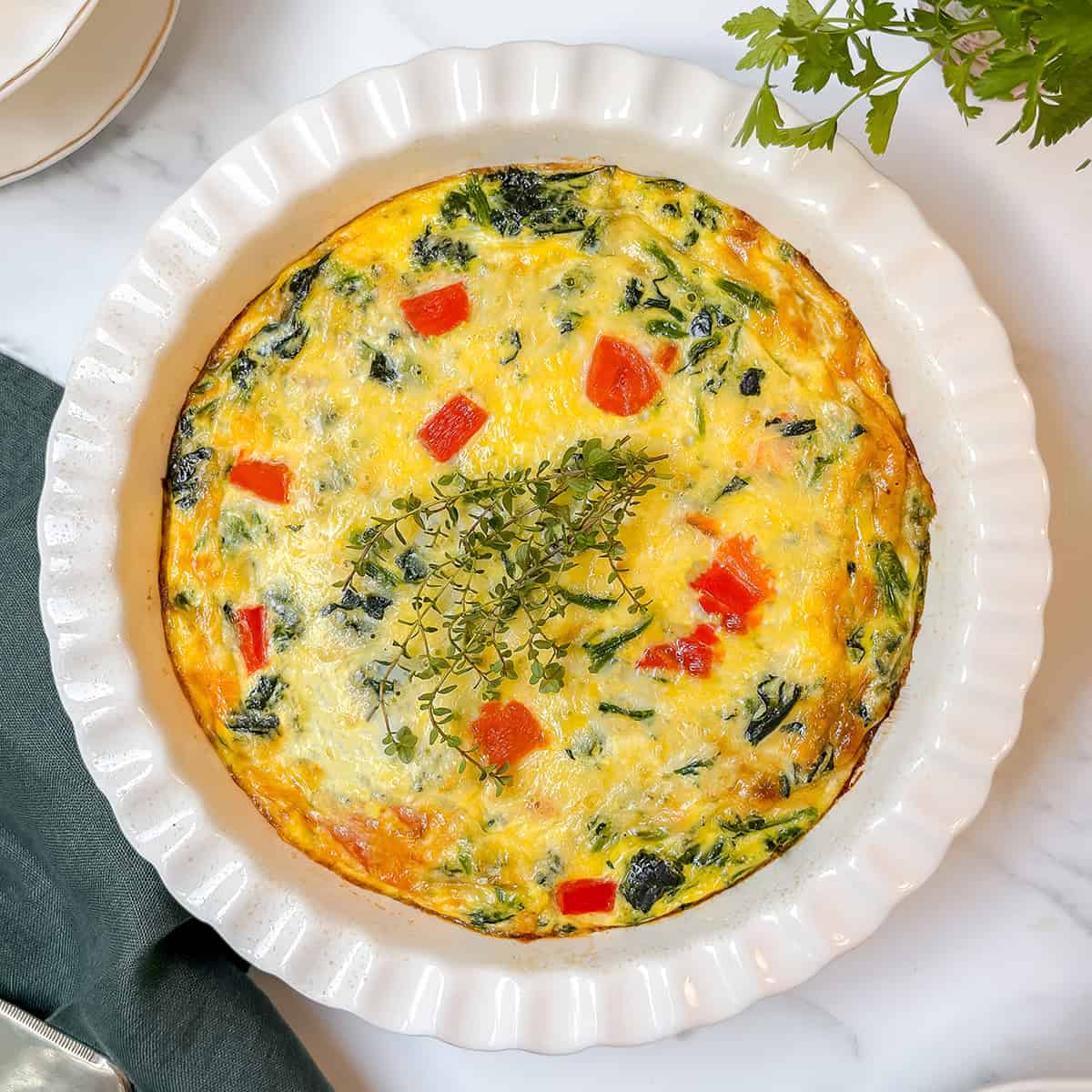 A baked vegetable frittata, perfect as a crustless quiche recipe, features red bell pepper pieces and leafy greens, garnished with fresh herbs, and sits in a white fluted dish on a marble surface.