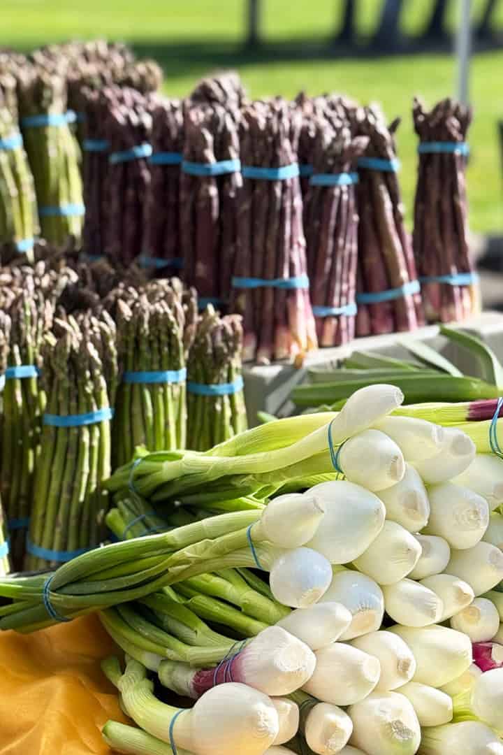 Bundles of fresh green and purple asparagus and white spring onions are displayed on a table at an outdoor market, perfect for adding a farm-fresh foods to 31Daily's Sunday Edit May Table.