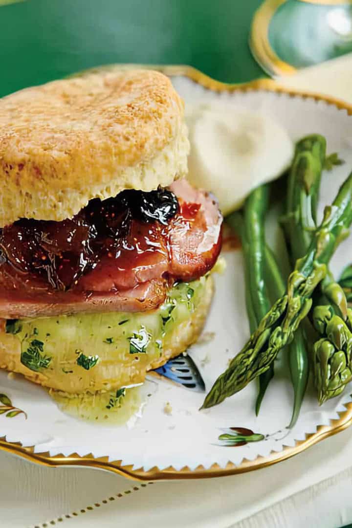 A plate with a biscuit sandwich filled with ham, cheese, and jam, served with steamed asparagus and green beans.