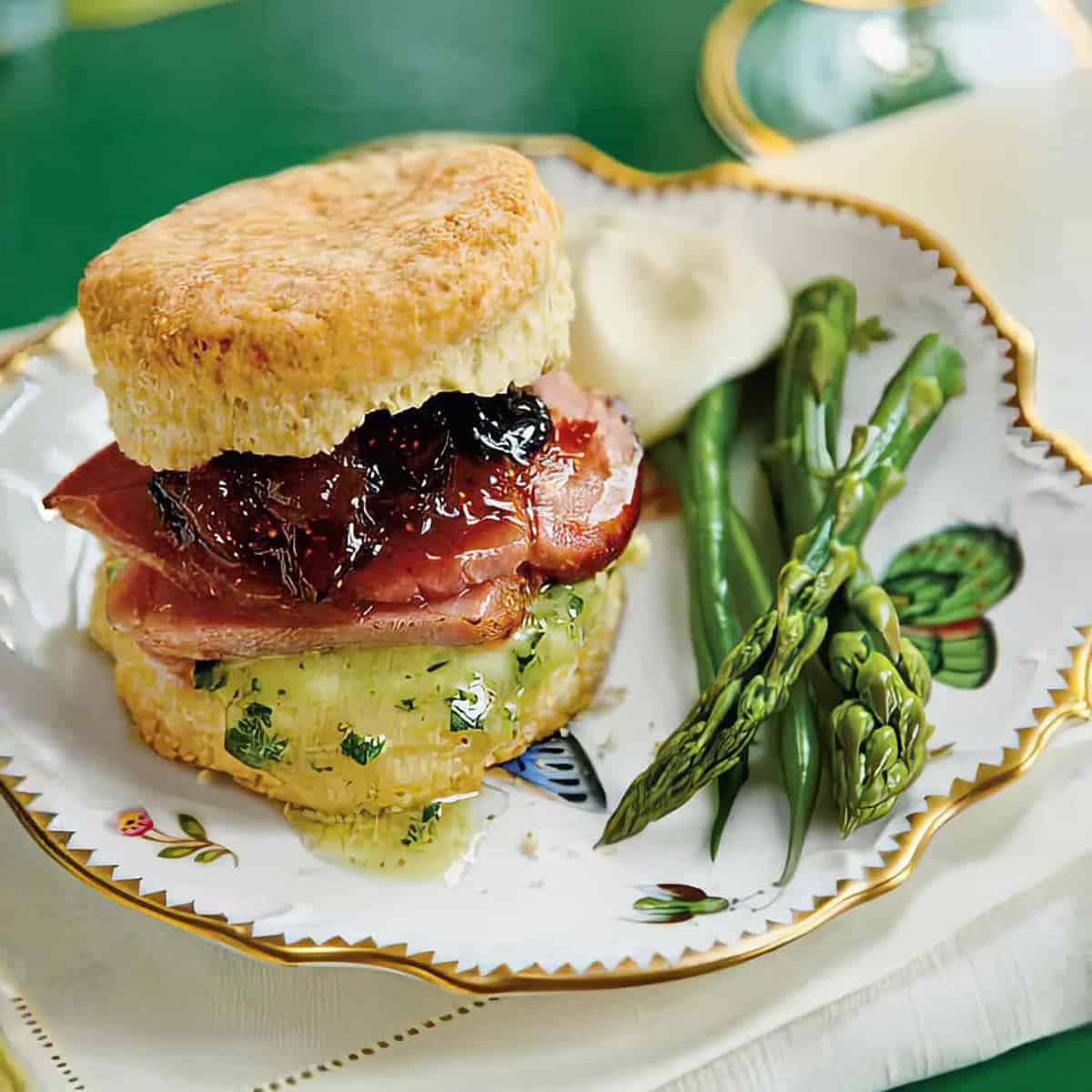 A plate with a biscuit sandwich filled with ham, cheese, and jam, served with steamed asparagus and green beans.