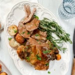 A plate with roast lamb, roasted potatoes, fresh rosemary, and gravy, garnished with chopped parsley on a white platter.