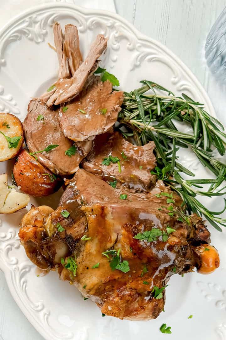 A plate with roast lamb, roasted potatoes, fresh rosemary, and gravy, garnished with chopped parsley on a white platter.