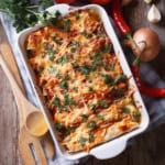 A baking dish filled with cheese-topped enchiladas garnished with fresh herbs, surrounded by wooden utensils, onions, chili peppers, and parsley on a wooden table.