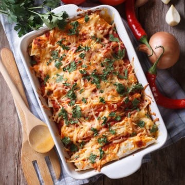 A baking dish filled with cheese-topped enchiladas garnished with fresh herbs, surrounded by wooden utensils, onions, chili peppers, and parsley on a wooden table.