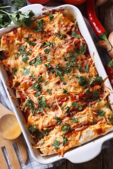 A baking dish filled with cheese-topped enchiladas garnished with fresh herbs, surrounded by wooden utensils, onions, chili peppers, and parsley on a wooden table.
