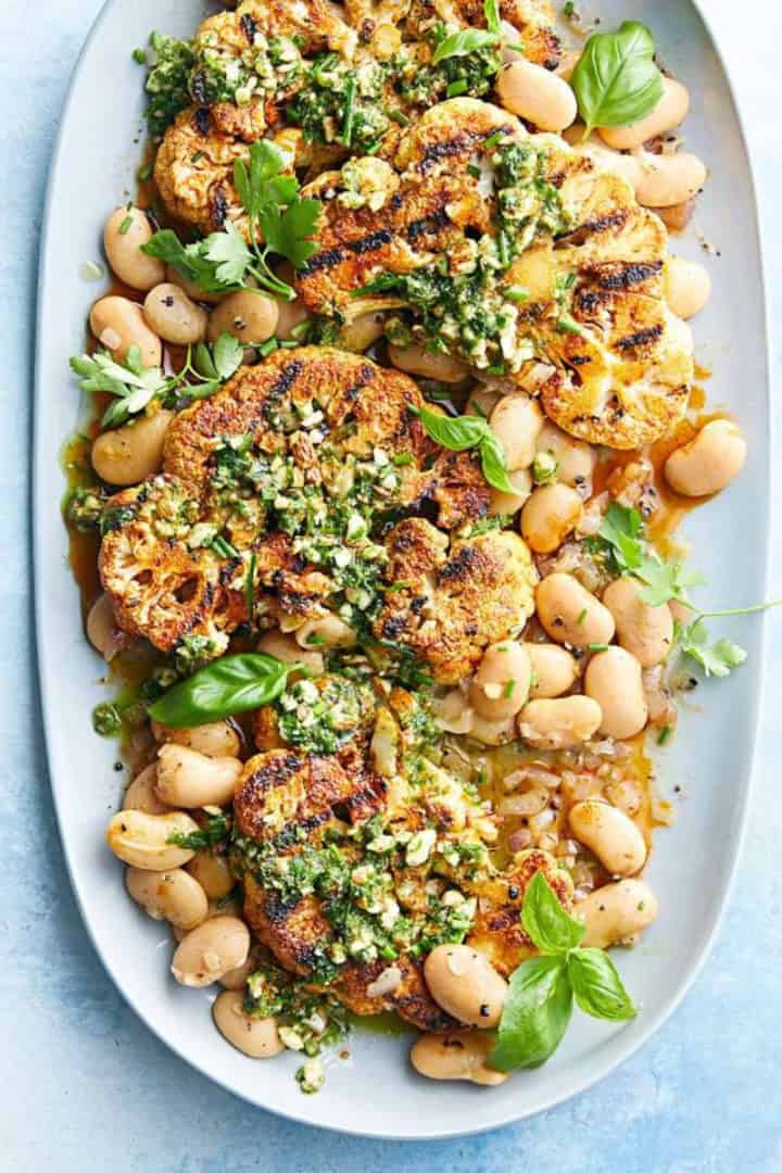 Grilled cauliflower steaks garnished with herbs and sauce, served on a platter with white beans and fresh basil leaves-perfect for light Spring Mediterranean Diet Dinners.