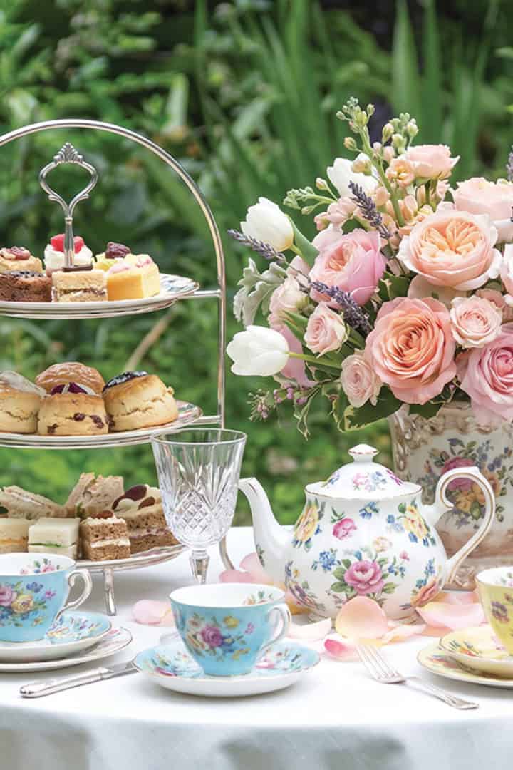 A table set for a Mother's Day Tea with floral china, a teapot, two cups, a tiered tray of pastries, and a bouquet of pink flowers, all beautifully arranged with greenery in the background.