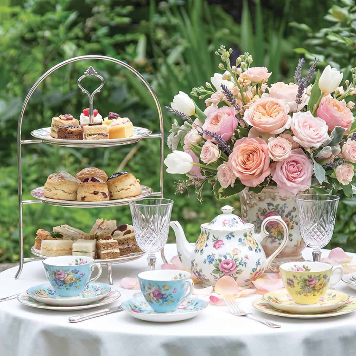A table set for a Mother's Day Tea with floral china, a teapot, two cups, a tiered tray of pastries, and a bouquet of pink flowers, all beautifully arranged with greenery in the background.