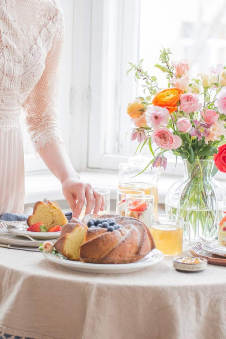 Serving full size desserts for afternoon tea at a table set with drinks and a vase of colorful flowers near a sunlit window.