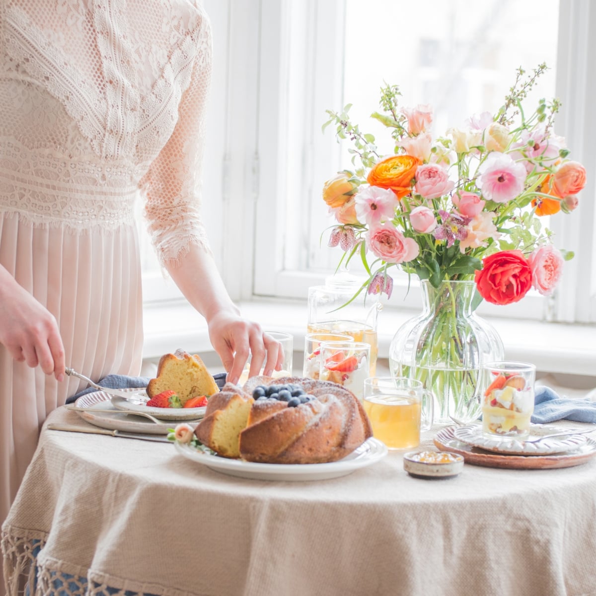 Serving full size desserts for afternoon tea at a table set with drinks and a vase of colorful flowers near a sunlit window.