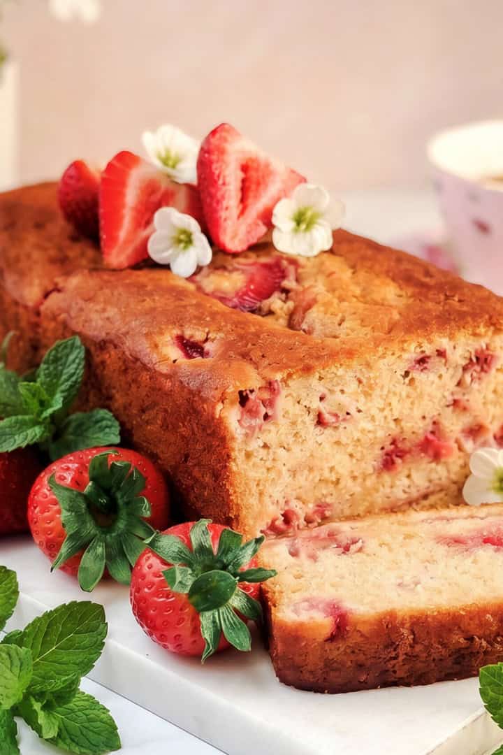A loaf of strawberry honey yogurt bread topped with fresh strawberries and white flowers, a slice cut, mint leaves beside it, and a cup of tea in the background.