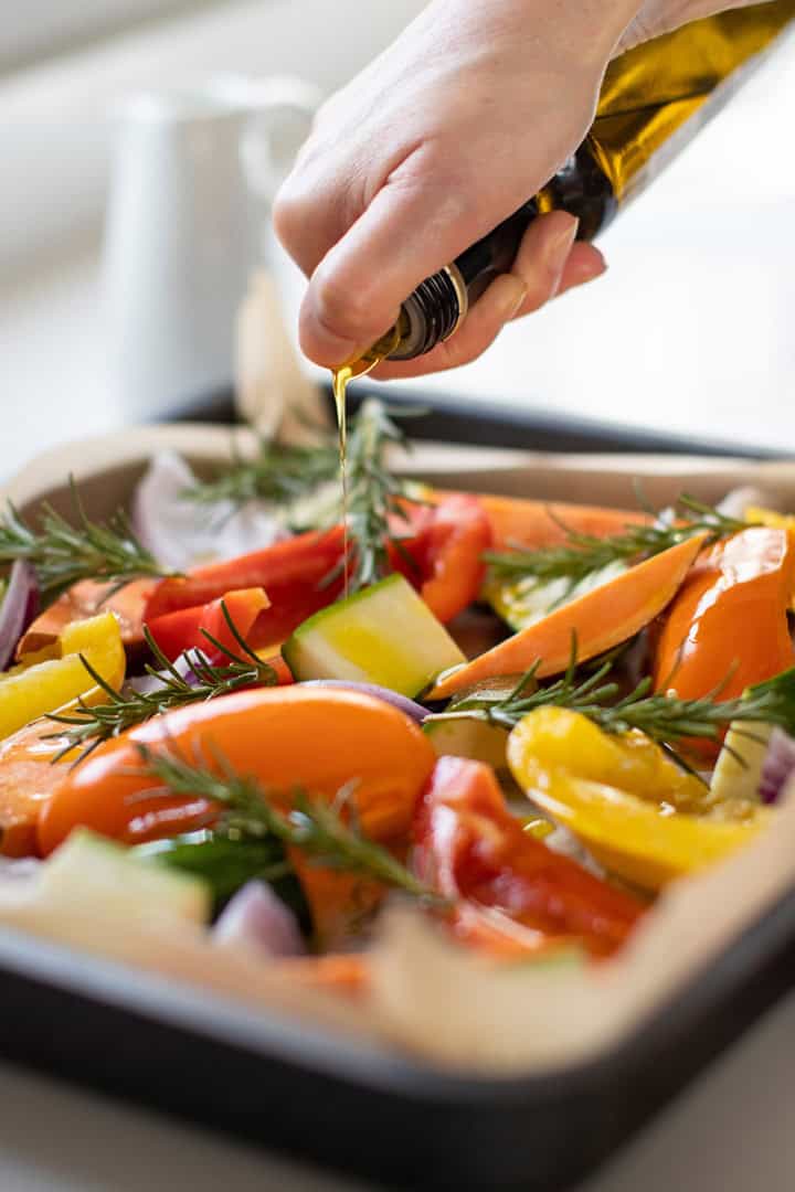 A hand drizzles olive oil over a tray of sliced vegetables and rosemary sprigs, preparing them for roasting-an inviting addition to any Easter table.