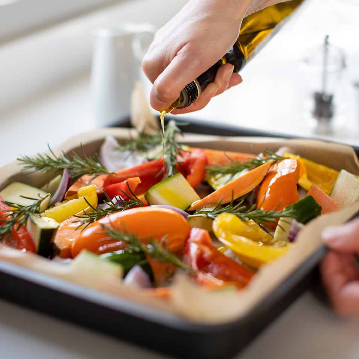 A hand drizzles olive oil over a tray of sliced vegetables and rosemary sprigs, preparing them for roasting-an inviting addition to any Easter table.