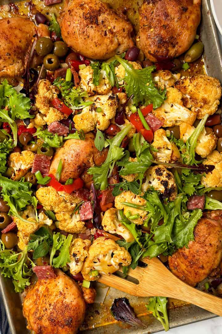 Sheet pan with baked chicken thighs inspired by Chicken Muffaletta, roasted cauliflower, olives, red bell peppers, leafy greens, and herbs, with a wooden spatula and blue-striped napkin on the side.