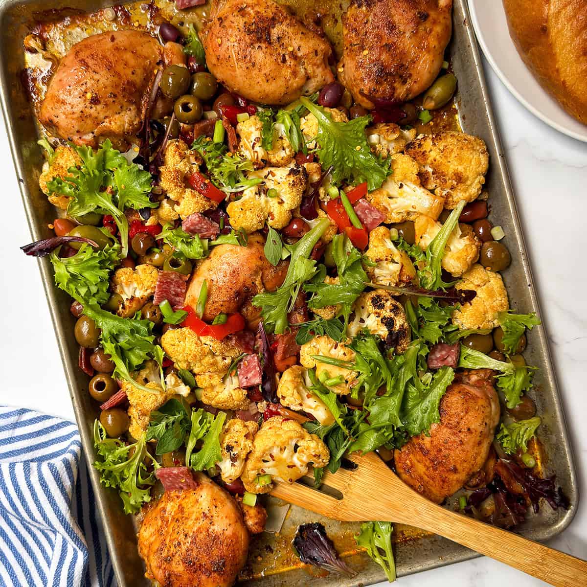 Sheet pan with baked chicken thighs inspired by Chicken Muffaletta, roasted cauliflower, olives, red bell peppers, leafy greens, and herbs, with a wooden spatula and blue-striped napkin on the side.