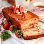 A loaf of strawberry yogurt bread with fresh strawberries and white flowers on top, one slice cut, displayed on a marble board with a teacup and striped napkin in the background. A delicious and healthy bread perfect for any occasion.