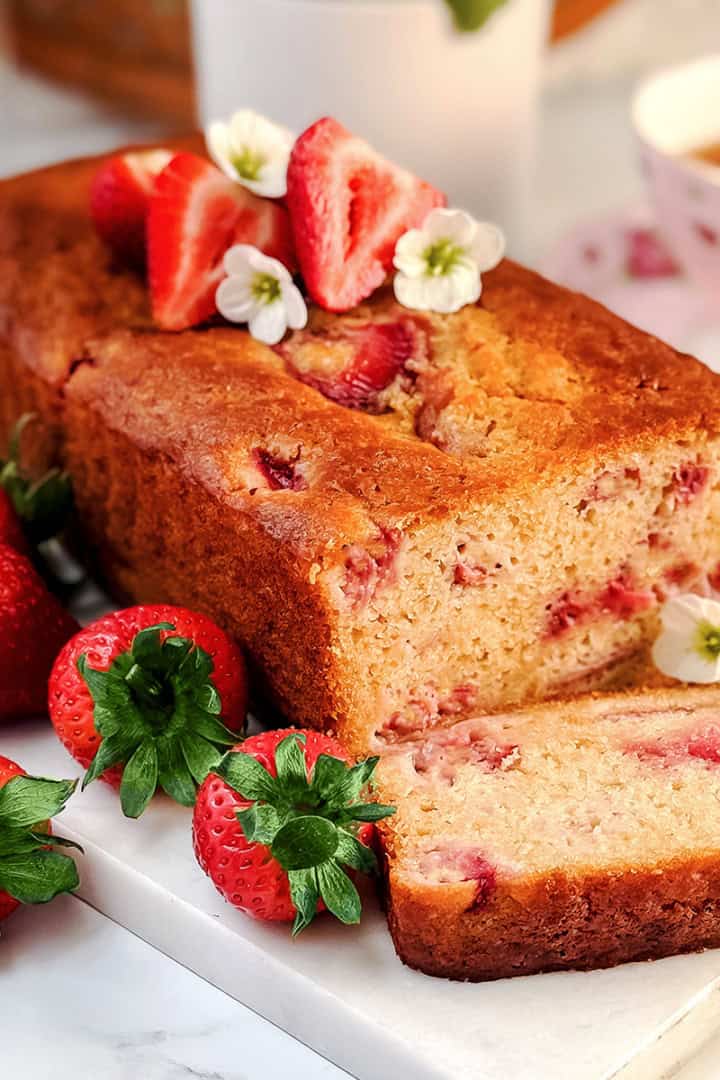 A loaf of strawberry yogurt bread with fresh strawberries and white flowers on top, one slice cut, displayed on a marble board with a teacup and striped napkin in the background. A delicious and healthy bread perfect for any occasion.