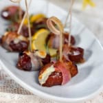 Blue cheese-stuffed dates wrapped in bacon, secured with toothpicks, make a savory appetizer, artfully arranged on a white platter with lemon slices in the background.