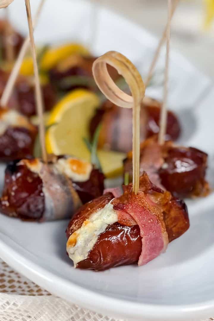 Blue cheese-stuffed dates wrapped in bacon, secured with toothpicks, make a savory appetizer, artfully arranged on a white platter with lemon slices in the background.