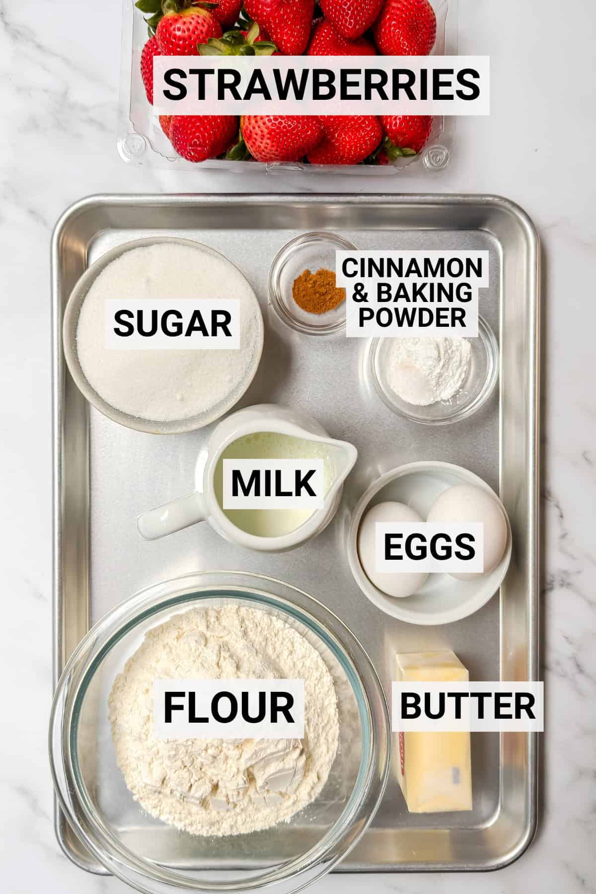 Tray with labeled ingredients for Strawberry Muffins: strawberries, sugar, cinnamon and baking powder, milk, eggs, flour, and butter on a marble surface.