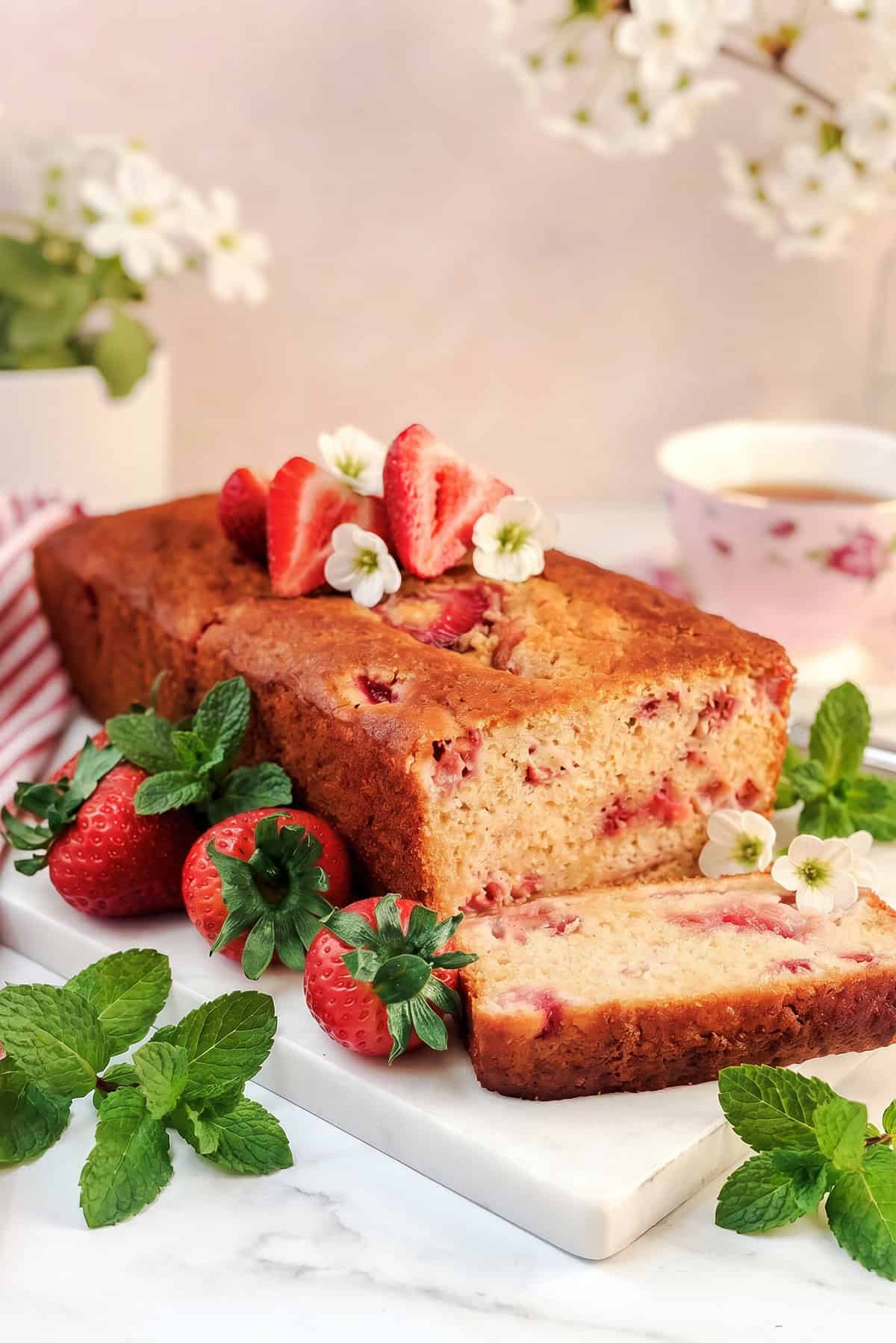 A sliced strawberry honey yogurt bread garnished with fresh strawberries, mint leaves, and white flowers on a marble board, with a teacup and flowers in the background.