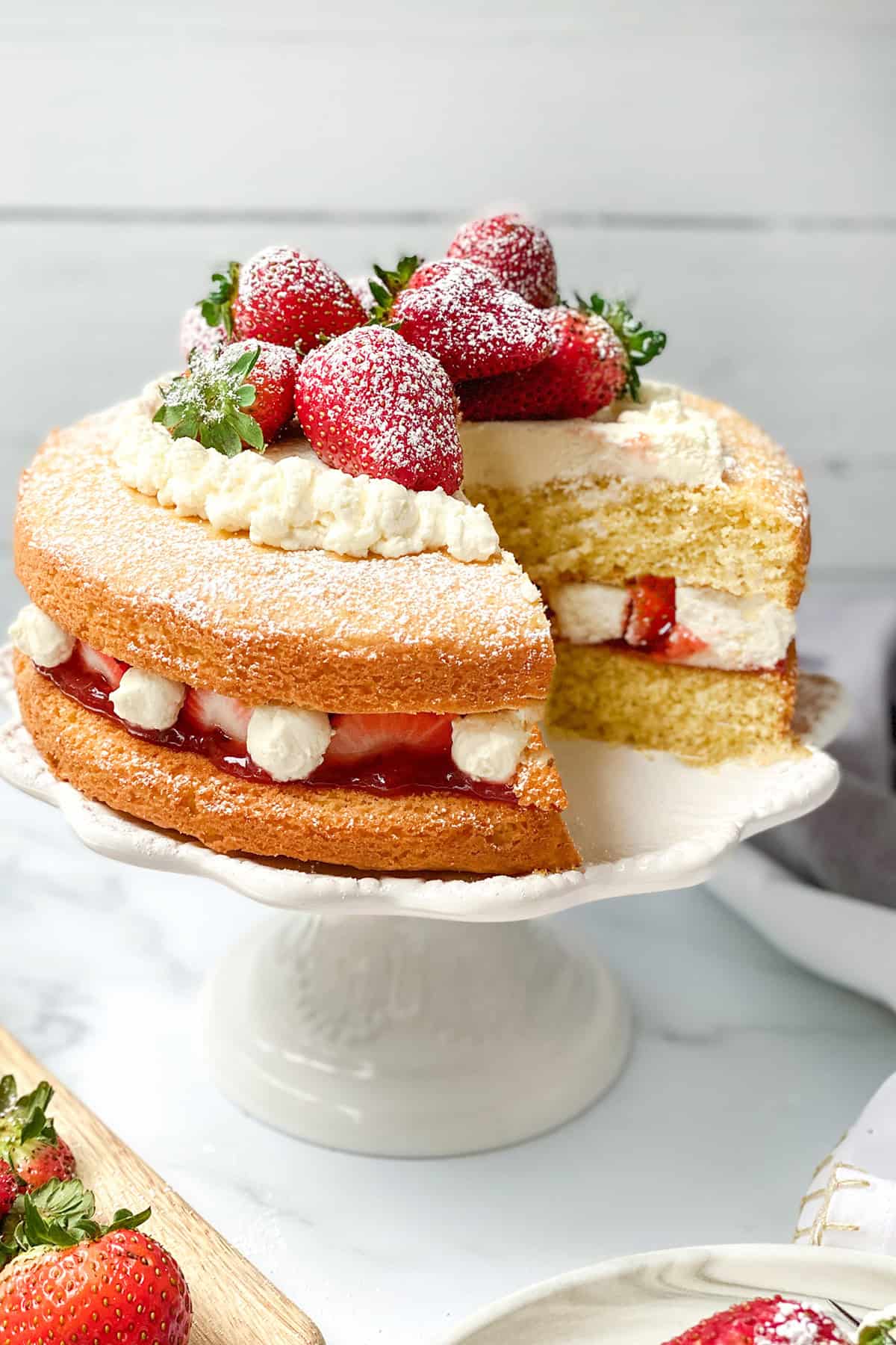 A layered sponge cake with cream and strawberries, topped with whole strawberries and powdered sugar, sits on a white cake stand with a slice removed-perfect among full size desserts for afternoon tea.