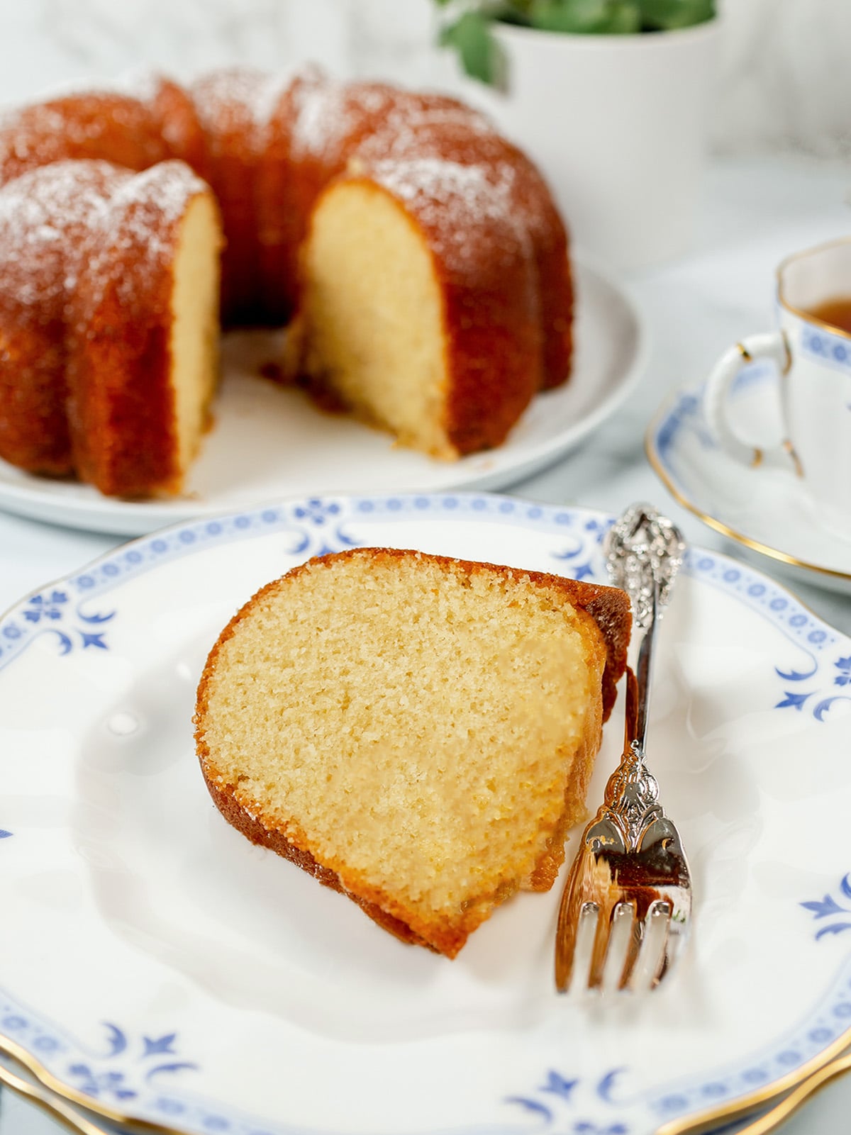 A slice of Kentucky Butter Cake on a decorative plate with a fork, the rest of the moist bundt cake in the background and a cup of tea on the side.