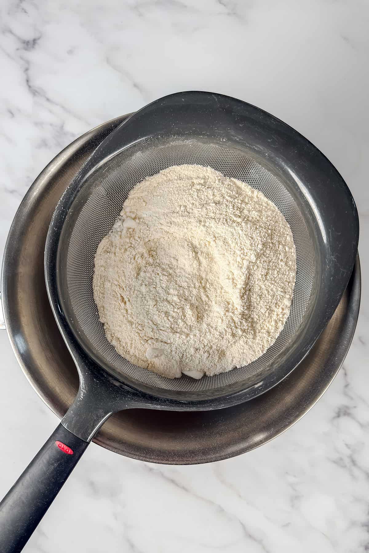 A metal sieve containing Kentucky Butter Cake ingredients rests over a stainless steel mixing bowl on a marble countertop.