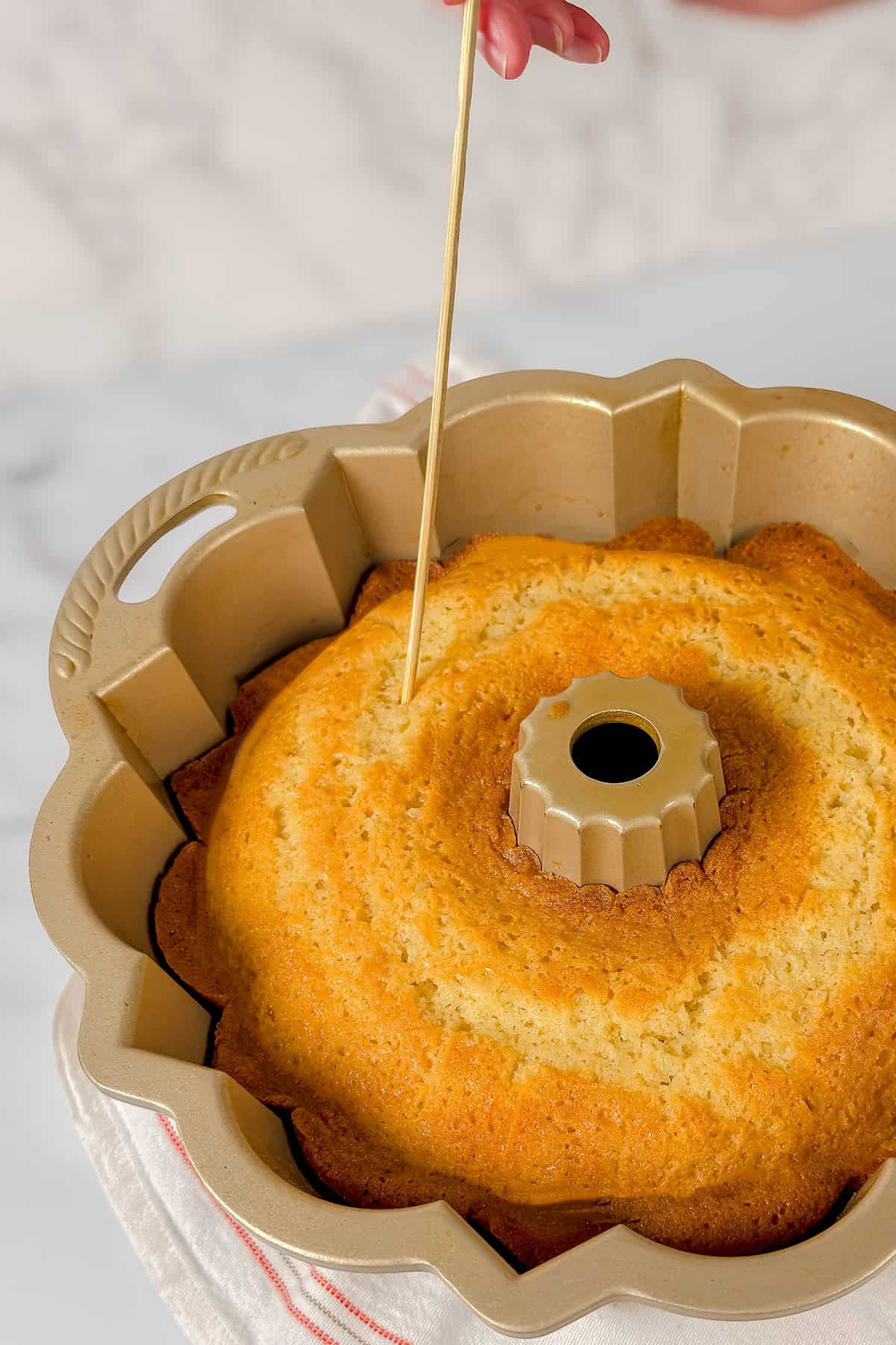 A hand inserts a wooden skewer into a golden-brown Kentucky Butter Cake in a Bundt pan preparing for the butter sauce. 