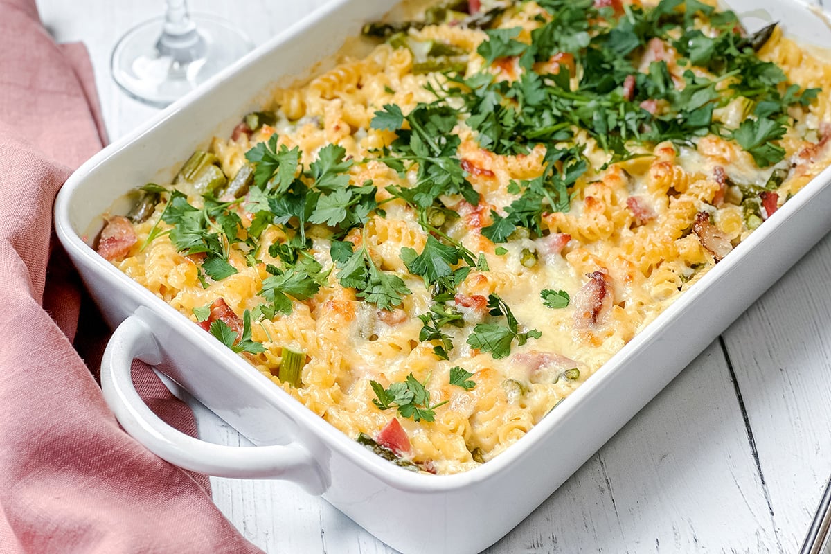 A baked pasta casserole with ham and tender asparagus, topped with melted cheese and chopped parsley, served as a main dish in a white rectangular dish on a white wooden table.