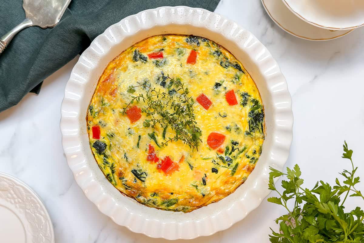 A baked vegetable frittata, inspired by a crustless quiche recipe, with red peppers and greens in a white ceramic dish, garnished with fresh herbs on a marble surface.