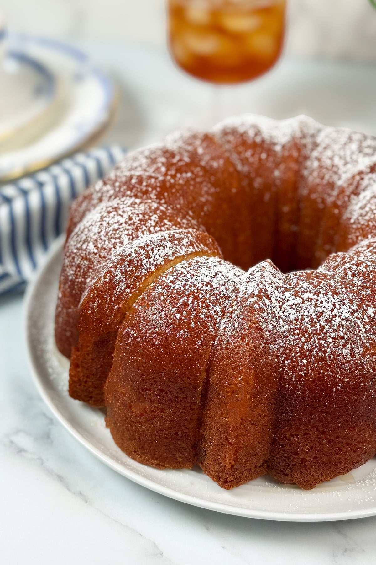 A golden brown Kentucky Butter Cake dusted with powdered sugar sits on a white plate atop a marble surface.