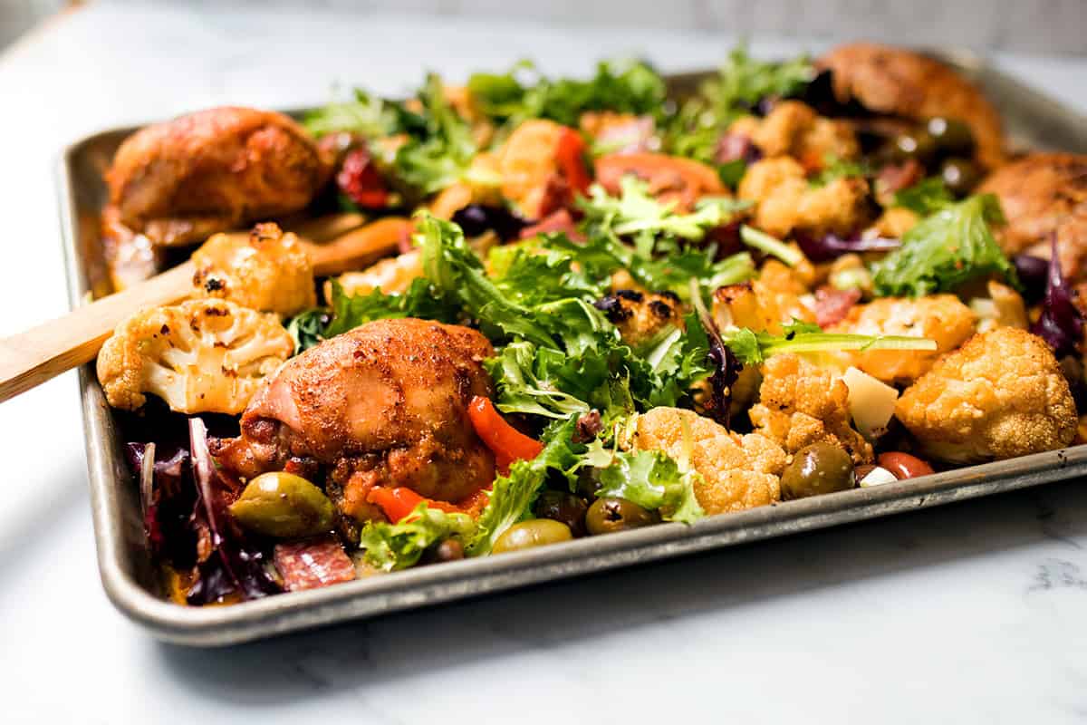 Sheet Pan Chicken Muffaletta with roasted chicken thighs, cauliflower, olives, greens, and red peppers on a white countertop.