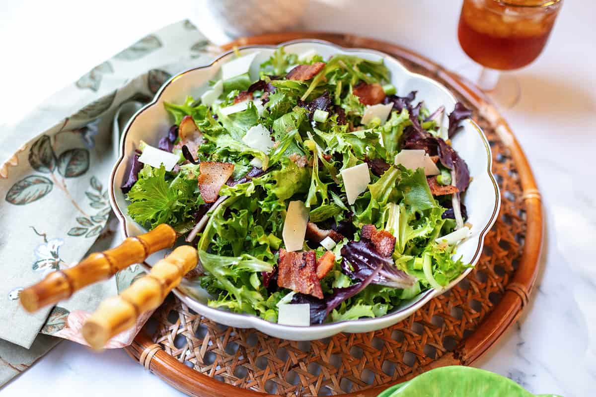 A bowl of spinach salad with bacon pieces, shaved cheese, and a drizzle of warm bacon dressing, served on a woven placemat with salad tongs and a napkin nearby.