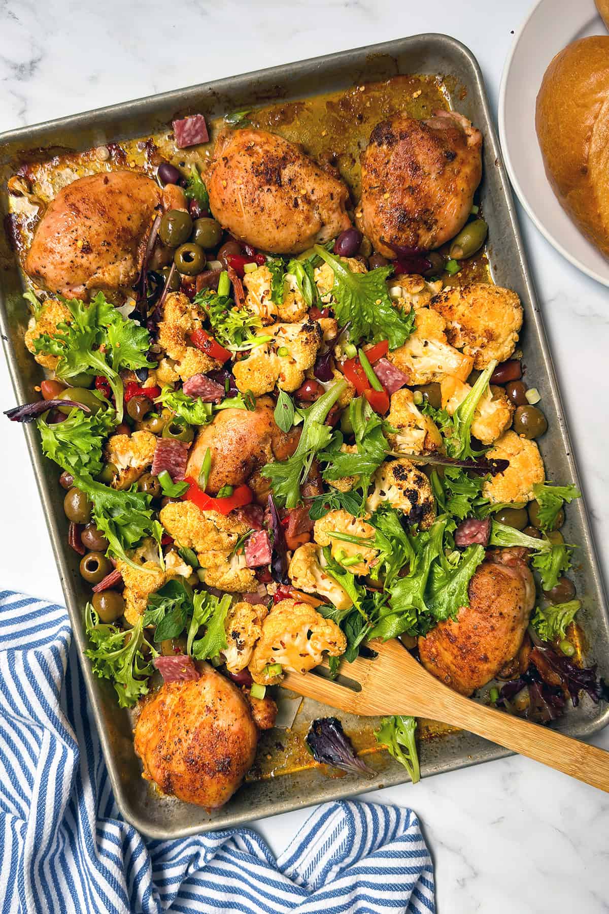 Sheet pan with roasted chicken thighs, cauliflower, olives, mixed greens, red peppers, and diced sausage&mdash;bringing muffaletta-inspired flavors to your table. Served on a marble surface with a wooden spoon and a bread roll nearby.