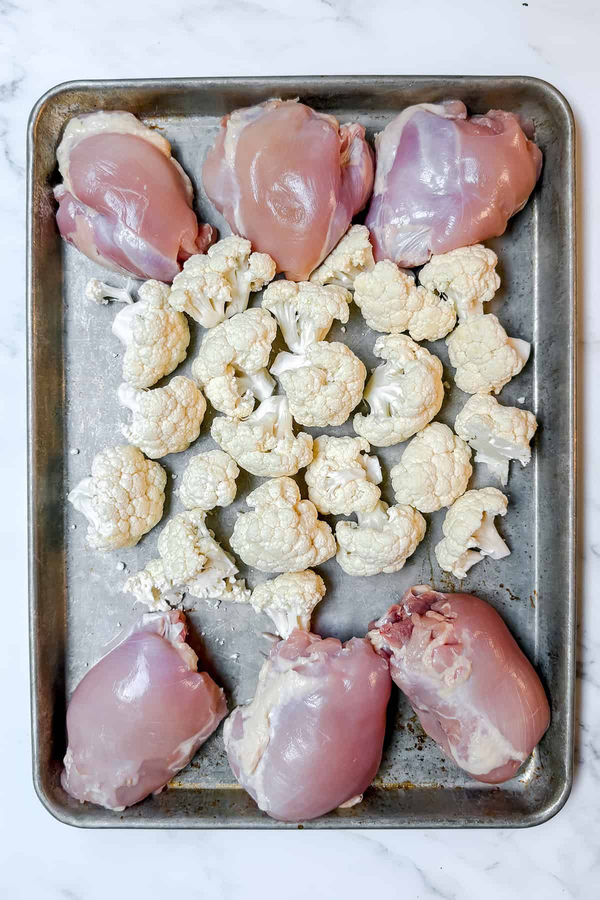 A sheet pan with raw chicken thighs arranged around the edge and cauliflower florets in the center, perfect for your next sheet pan chicken dinner.
