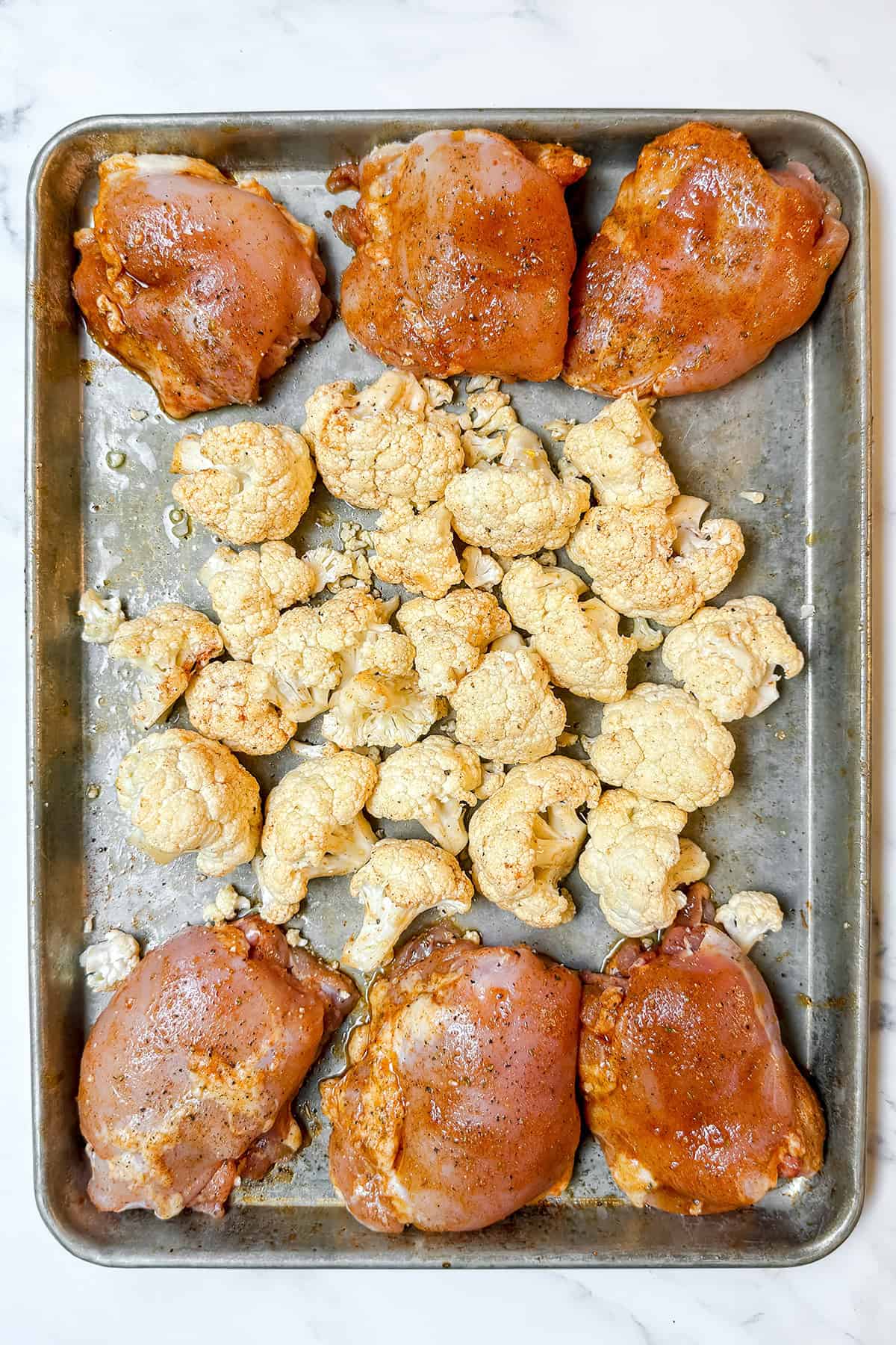 Raw, seasoned chicken thighs and cauliflower florets are arranged on a sheet pan, ready for roasting on a white marble surface.