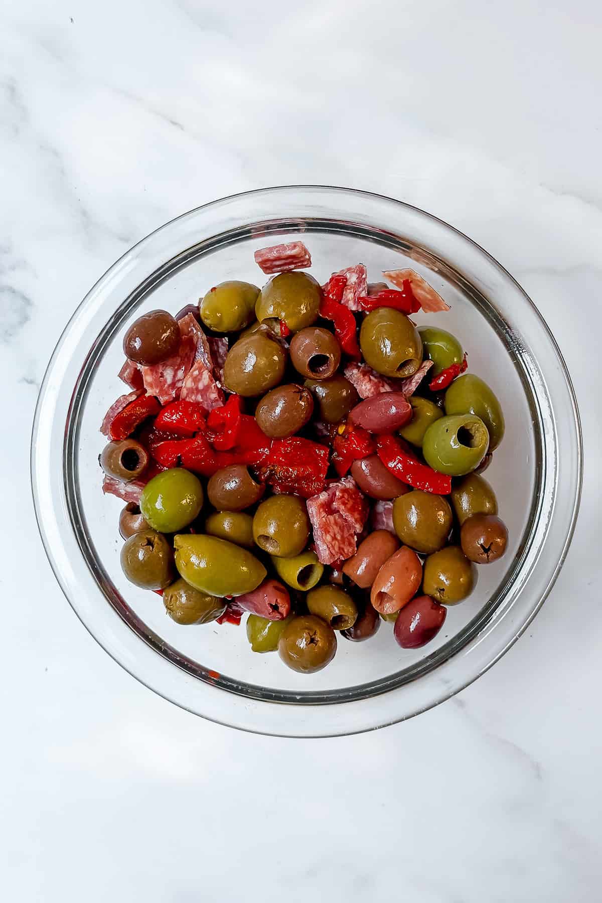 A glass bowl filled with assorted green and black olives, sliced red peppers, pieces of cured meat, and classic muffaletta flavors sits on a white marble surface.