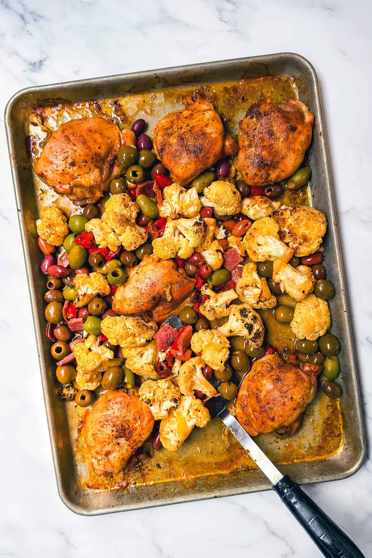 A sheet pan with roasted chicken thighs, cauliflower florets, green and black olives, and red peppers sits on a marble surface. A serving knife rests on the pan-perfect for an easy Sheet Pan Chicken muffaletta-inspired dinner.