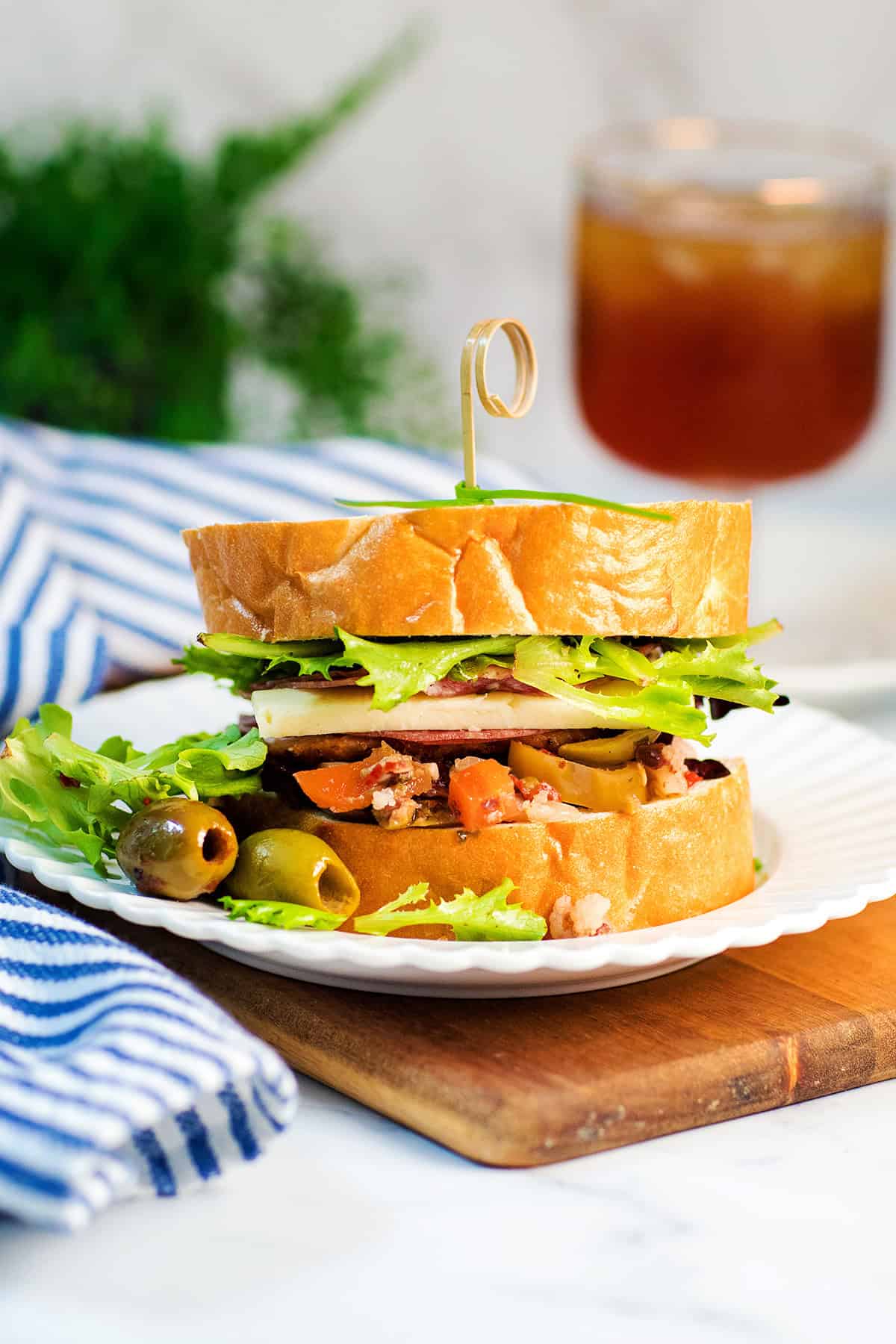 A Chicken Muffaletta sandwich with lettuce, cheese, and assorted vegetables sits on a white plate with two green olives, next to a striped napkin and a drink in the background.