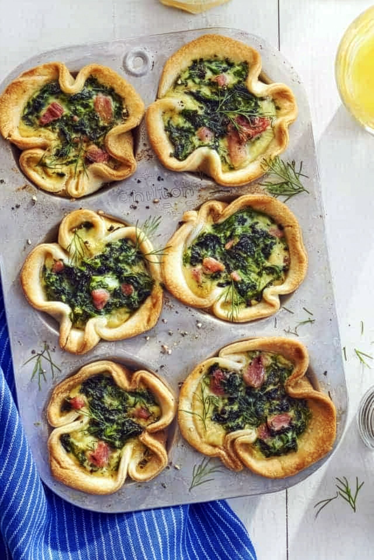A muffin tin with six ham and egg bakes filled with spinach, herbs, and diced ham sits on a white surface next to a blue striped cloth for a Mother's Day Brunch.