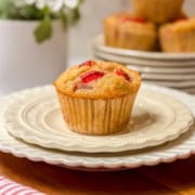 A single strawberry muffin sits atop a stack of white plates, with more delicious strawberry muffins and a potted plant softly blurred in the background.